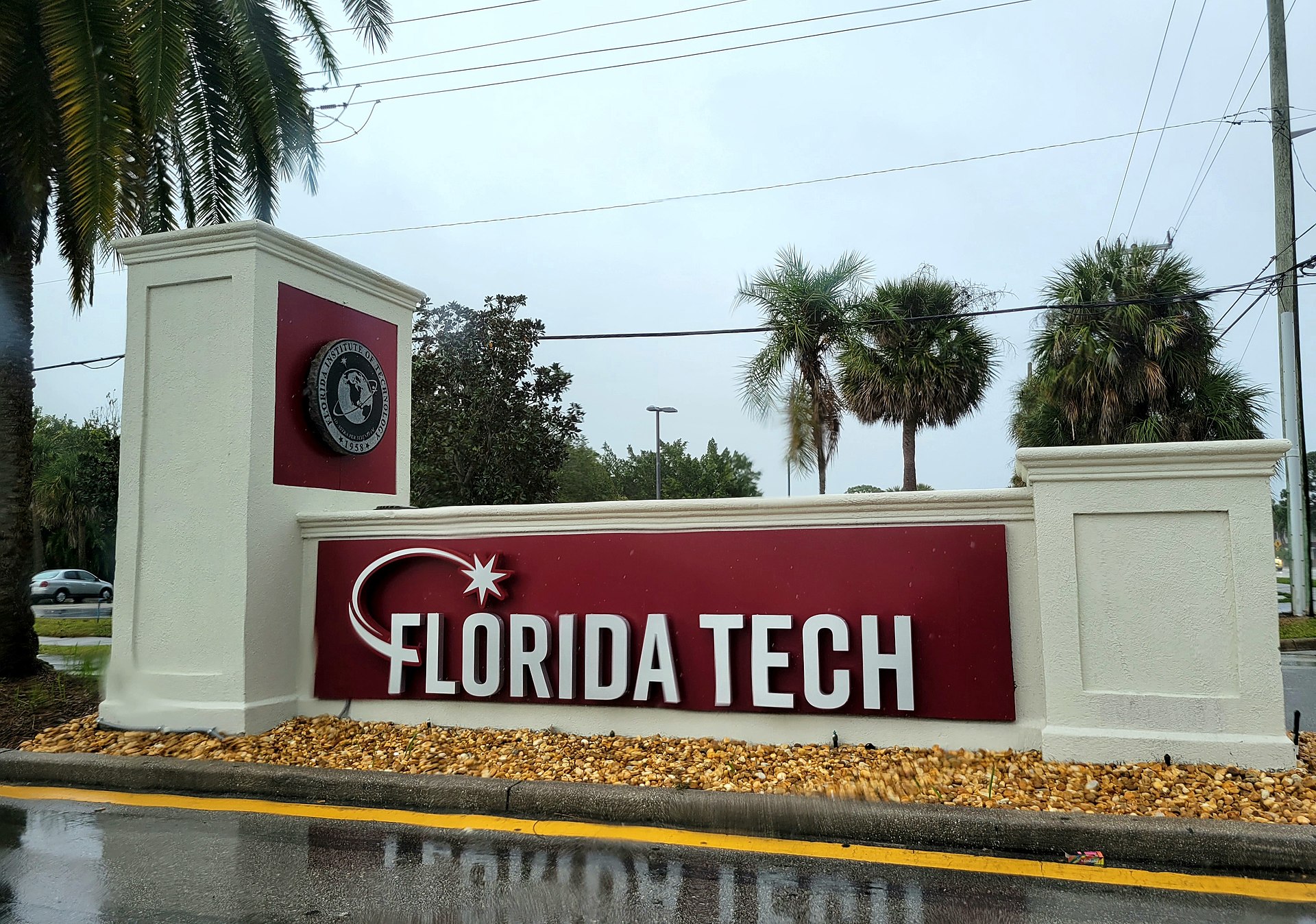 The Florida Institute of Technology entrance sign on a campus road in Melbourne, Florida, with palm trees and the campus visible beyond.